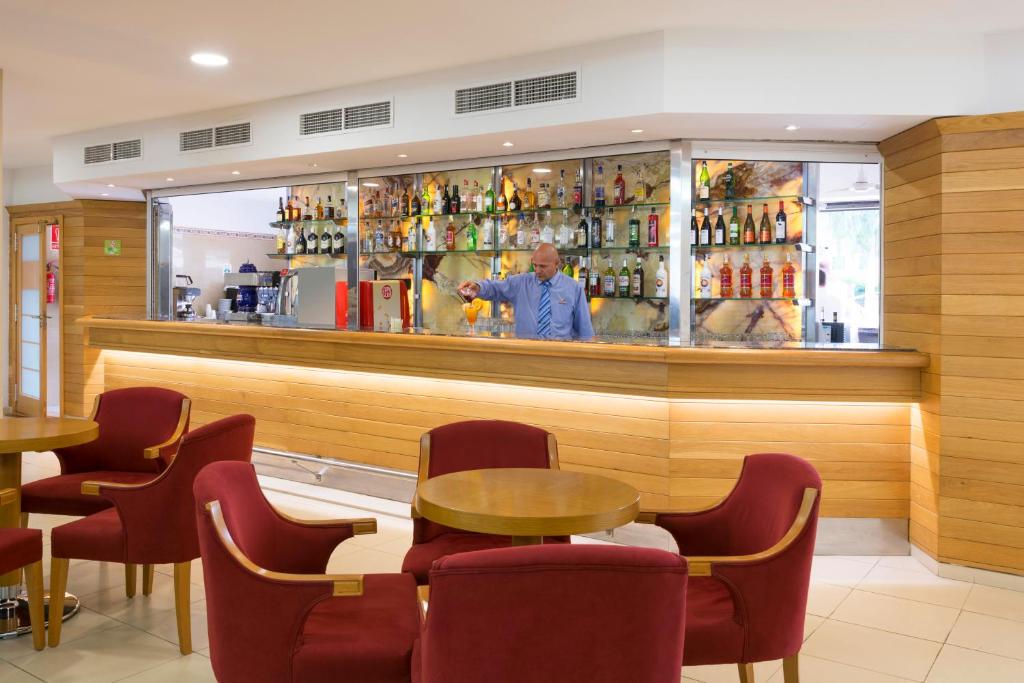 Bāra skats ar apkalpojošo darbinieku, kurš piedāvā dzērienus, un pie bāra ir sarkanās krēsli un koka galdi. Interjers ir mūsdienīgs un gaumīgs, radot patīkamu atmosfēru.