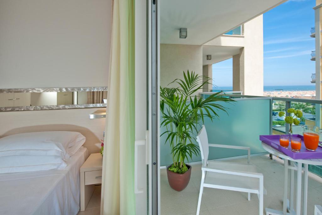 Mūsdienīgs viesnīcas numurs ar balkonu, kurā redzams jūras skats. Uz galda atrodas dzērieni un ziedi, bet istabā ir ērta gulta un mēbelēta telpa.