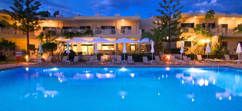 Vasaras nakts skats uz viesnīcu ar baseinu, apgaismojot ūdeni zilā krāsā un ainavu ar palmām un sauļošanās krēsliem.