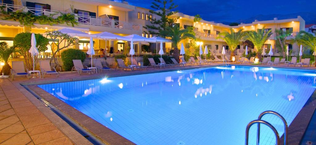 Lielisks viesnīcas baseins ar zilu ūdeni un ērtām sauļošanās vietām, apgaismots vakara stundās. Apkārt baseinam ir zaļumi un palmas, radot relaksējošu atmosfēru.