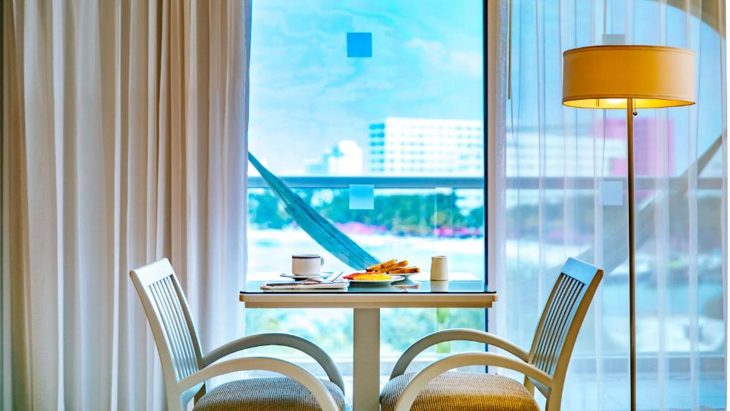 Gaiša istaba ar galdu un divām krēslām, uz galda ir brokastu ēdieni un dzērieni, logā redzams skats uz ārpusi.