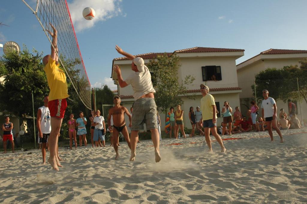 Pludmales volejbola spēle ar spēlētājiem, kas izpilda uzbrukumu, un skatītājiem, kas vēro. Apspēlēšana notiek saulainā dienā pludmalē, ar mājām fonā.