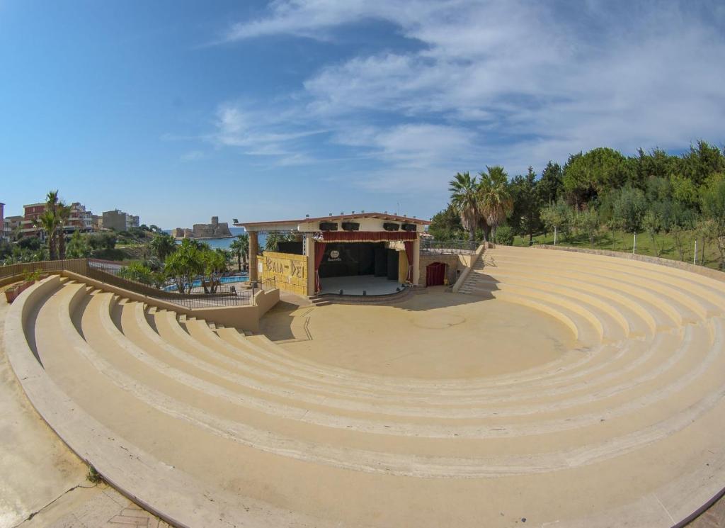 Atvērta amfiteātra skatuve ar soliņiem un skatu uz jūru, ieskauta palmu un zaļumu. Ideāla vieta kultūras pasākumiem un izklaidei.