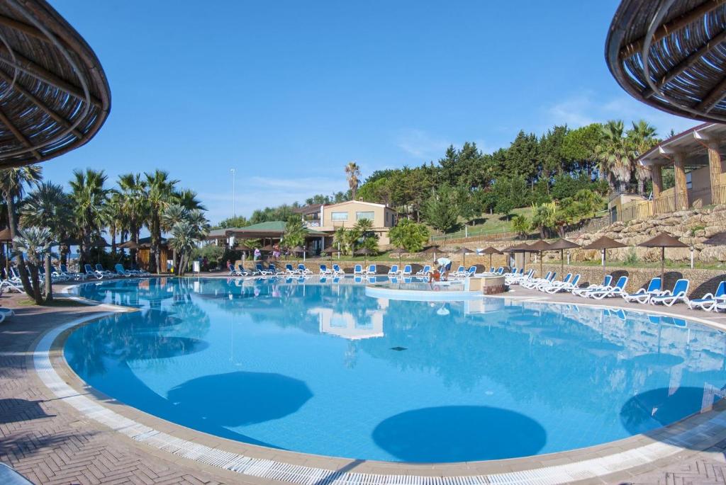 Lieliska peldbaseina ar palmām un sauļošanās krēsliem, kas piedāvā atpūtu saulainā vidē. Fonā redzami zaļi kalni un atpūtas zonas.