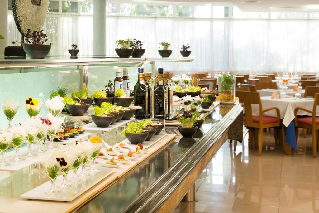 Eleganta bufete ar dažādiem salātiem un uzkodām, dekorēta ar zaļumiem un ziediem, fons ir gaiša restorāna telpa ar ērtām mēbelēm.