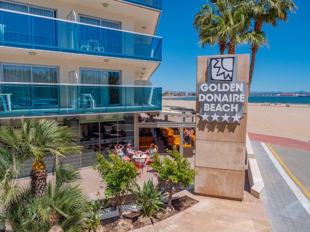 Zelta Donaire pludmales viesnīcas eksterjērs ar palmām un saulainu pludmali fonā. Viesi atpūšas terasē, baudot brīvdienu atmosfēru.