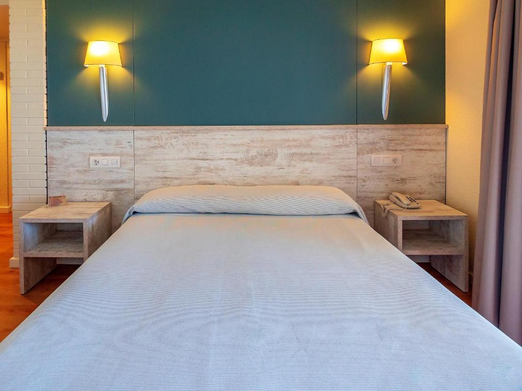 Mūsdienīgs guļamistabas interjers ar gultu, koka naktsgaldiņiem un sieniņu apgaismojumu, radot mierīgu atmosfēru. Gaišas krāsas un minimālisma dizains veicina relaksāciju.