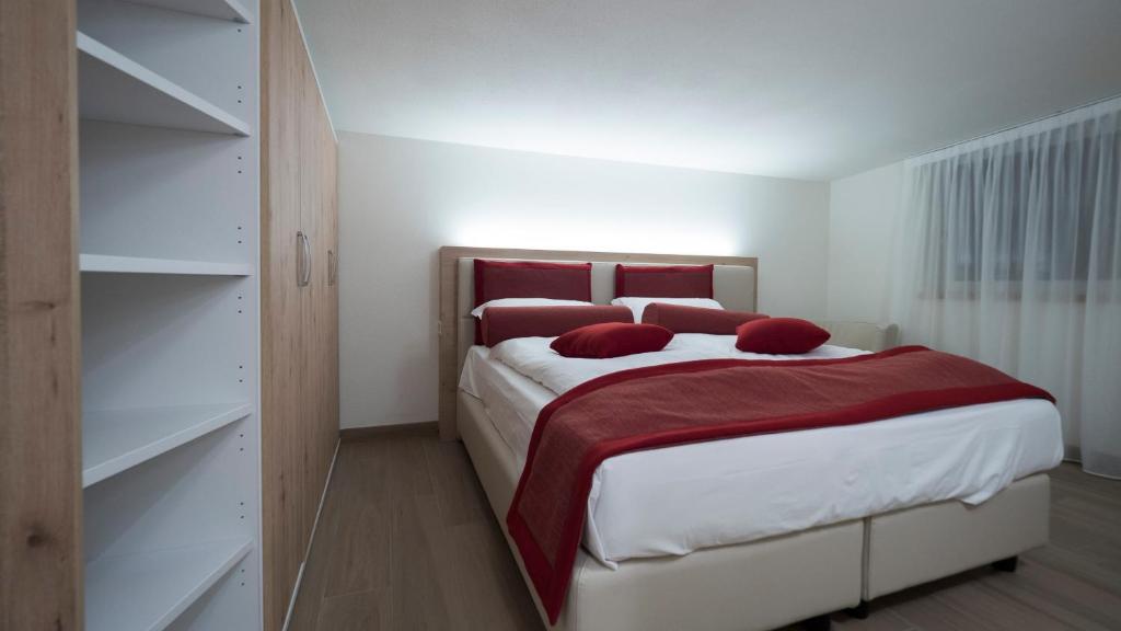 Mūsdienīgs guļamistabas interjers ar plašu gultu, sarkanām spilveniem un koka mēbelēm, radot mājīgu atmosfēru. Gaisma un interjers veido elegantu un relaksējošu vidi.