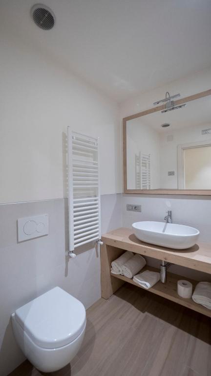 Mūsdienīgs vannas istabas interjers ar minimālistisku dizainu, balto izlietni un koka plauktu. Redzams arī tualete un apsildāms dvieļu žāvētājs.