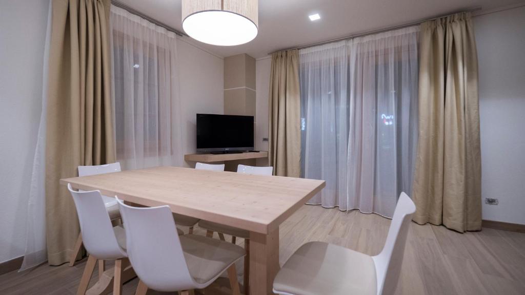 Mūsdienīga viesistaba ar koka galdu un baltām krēslām, gaišām aizkariem un televizoru. Interjers ir vienkāršs un elegants, radot mājīgu atmosfēru.