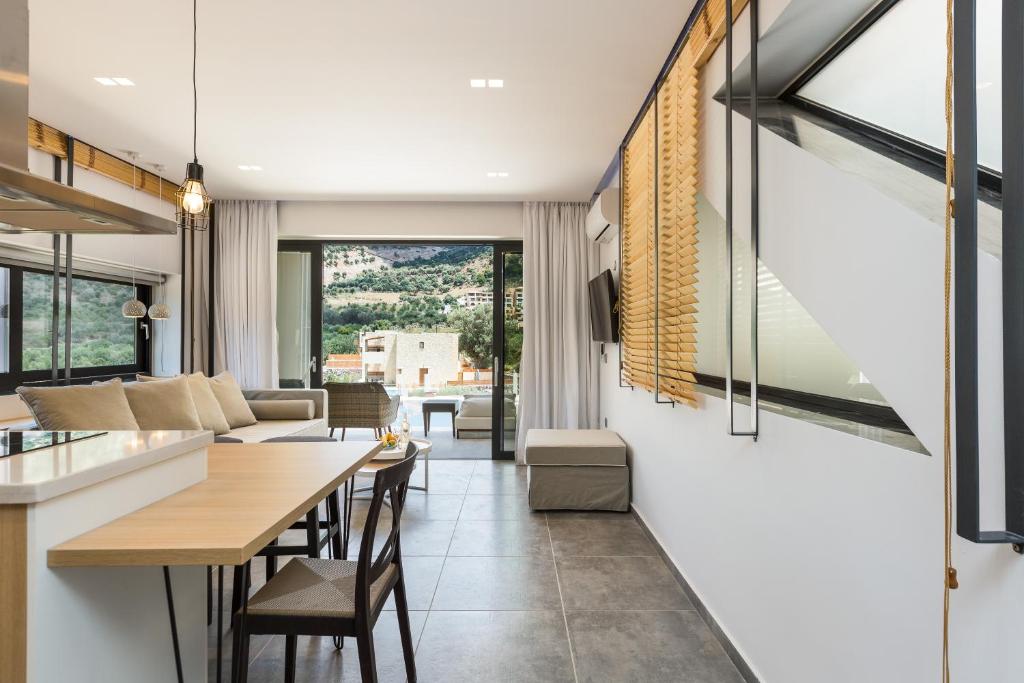 Mūsdienīgs interjers ar atvērtu plānojumu, kas ietver ēdamgaldu un viesistabu ar logiem, caur kuriem redzams ainava. Interjerā ir gaišas krāsas un minimālistiski elementi.