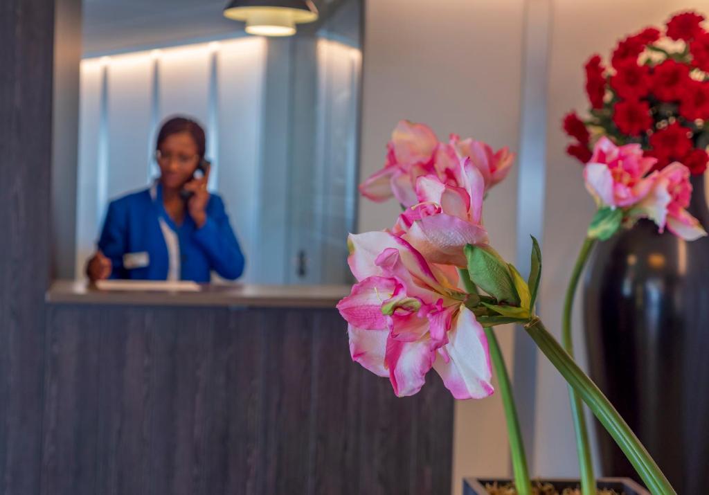 Reģistratūras darbiniece, kas runā pa telefonu, ir fona plānā, kamēr priekšplānā ir ziedi ar rozā un baltiem ziediem. Svaigas puķes piešķir viesnīcas iekārtojumam eleganci un siltumu.
