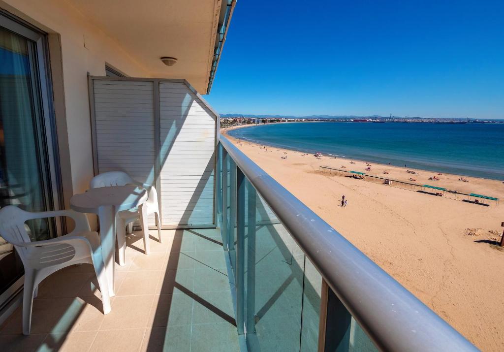 Balkons ar skatu uz pludmali, kurā redzama zila jūra un smilšu krasts. Pie galda ir divas baltas plastmasas krēsli.