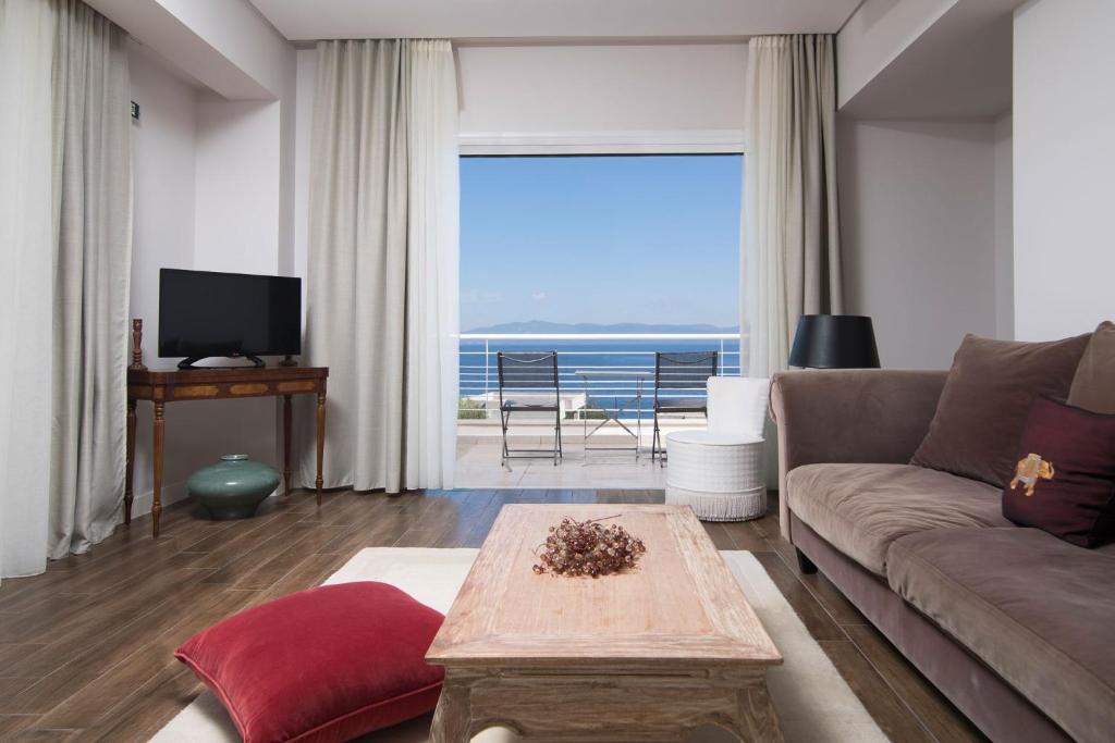 Mūsdienīga viesistaba ar skatu uz jūru, kurā ir dīvāns, televizors un koka galdiņš. Telpā ir gaiši aizkari un eleganta interjera elementi, radot relaksējošu atmosfēru.