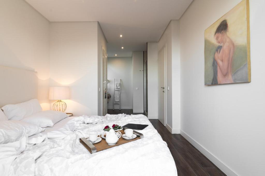 Mūsdienīgs guļamistabas interjers ar gultu, kurā ir balti gultas veļas audumi un koka paliktnis, uz kura ir tējkanna un krūzes. Sienā ir gleznas, kas piešķir telpai māksliniecisku noskaņu.