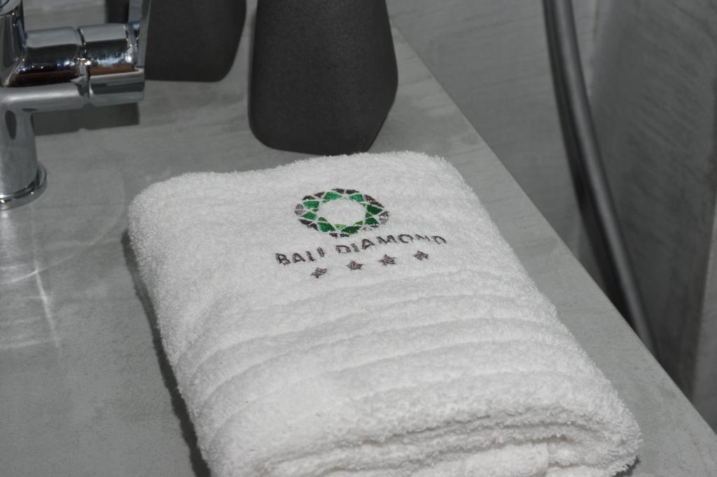 Balts dvielis ar "Bali Diamond" logo, novietots uz pelēka galda. Dvielis ir mīksts un izskatās viesnīcā.