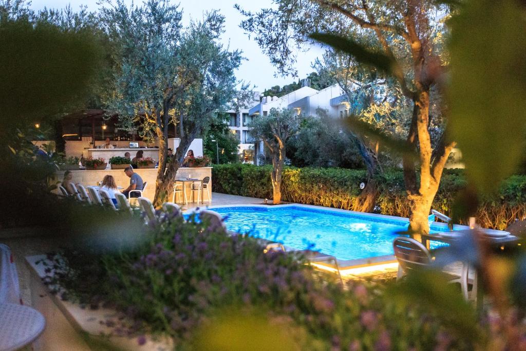 Nakts ainava ar peldbaseinu, apzaļumotu teritoriju un atpūtas zonu, kur cilvēki bauda vakaru. Ap gaismu apgaismotas olīvkoki un ziedi piešķir romantisku atmosfēru.