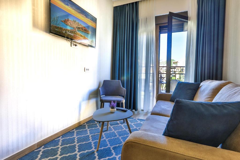 Mūsdienīgs viesistabas interjers ar dīvānu, TV un logu, caur kuru redzams balkons. Svaiga un gaisīga atmosfēra ar zilu paklāju un dekoratīvām spilveniem.