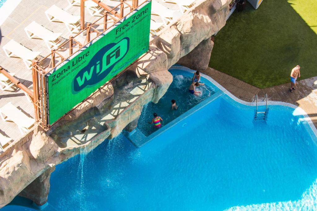 Peldbaseins ar brīvdabas reklāmu par bezvadu internetu, kur bērni un pieaugušie izbauda ūdens aktivitātes. Apkārt peldbaseinam ir saules gultas un zaļā zāle.