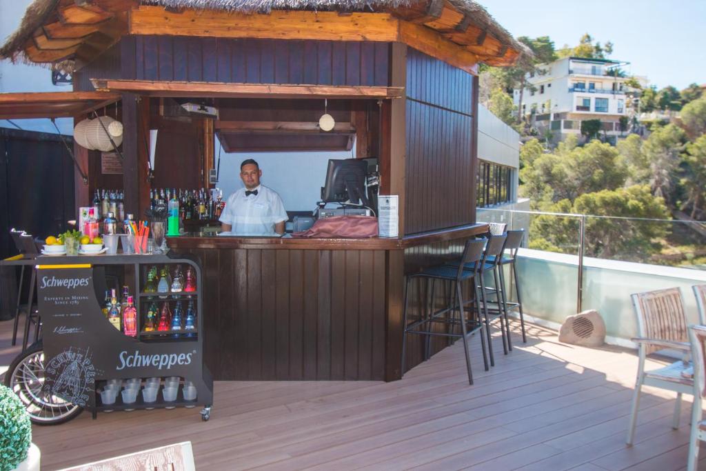 Bāra skats ar apkalpojošu darbinieku, kas stāv aiz koka bāra letes. Fons ietver dzērienu plauktus un mūsdienīgu ēku, kas atrodas blakus. Ideāla vieta atpūtai un izklaidei.