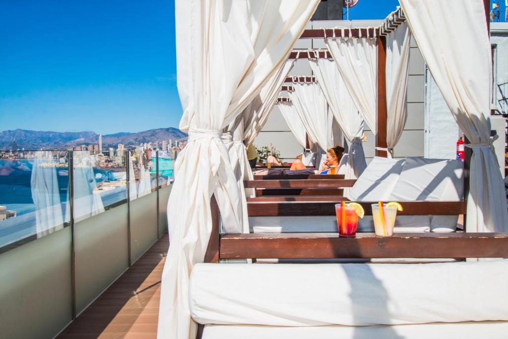 Lieliska skatu terase ar šūpuļtīklu, kurā cilvēki atpūšas, baudot kokteiļus un saules starus. Fons ir pilsētas ainava ar kalniem un jūru.