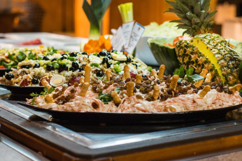 Krāsains bufete ar dažādiem salātiem un uzkodām, tostarp garnelēm un dārzeņiem, ar ananāsu un melone fonā.