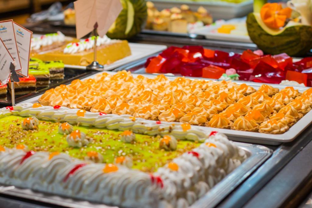 Saldumu bufete ar dažādiem krāsainiem desertiem, tostarp kūkām, želejām un krēmveida saldumiem, kas rotāti ar augļiem un riekstiem.