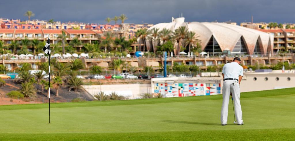 Golfista spēle uz zaļā laukuma ar mūsdienīgu ēku un palmām fona. Ideāla ainava golfa entuziastiem.