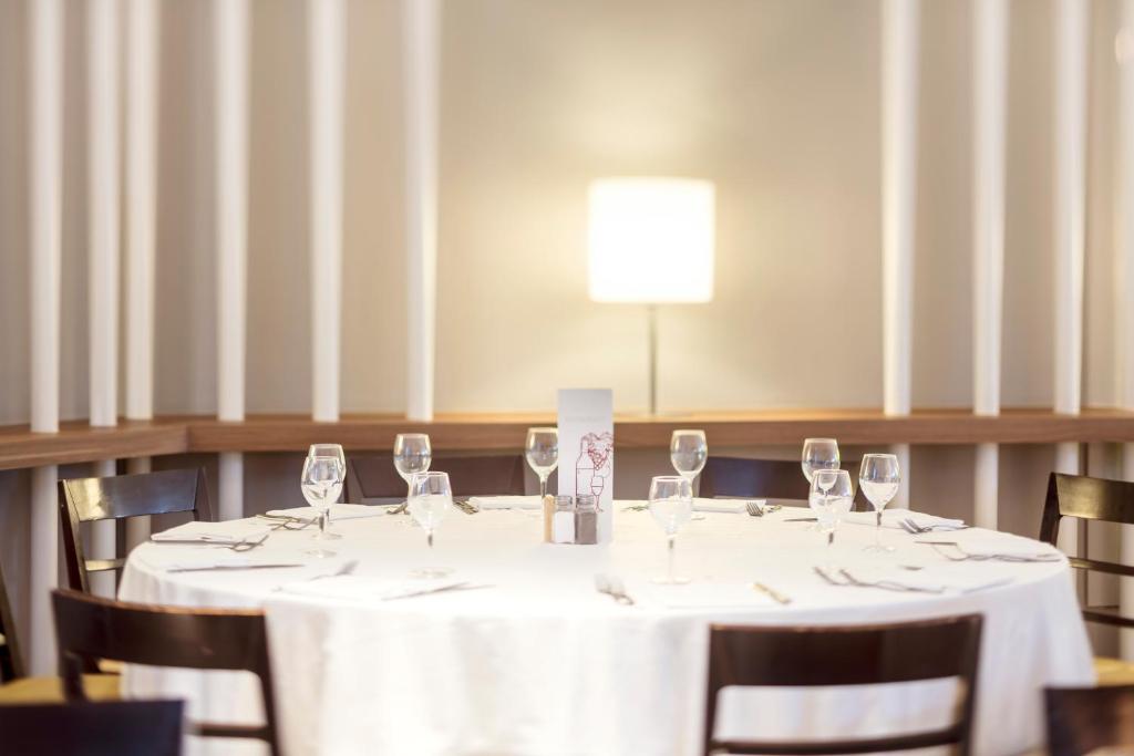 Eleganti noformēts pusdienu galds ar baltu galdautu un glāzēm, apgaismots ar mūsdienīgu lampu. Galdā ir ēdienkarte un galda piederumi.