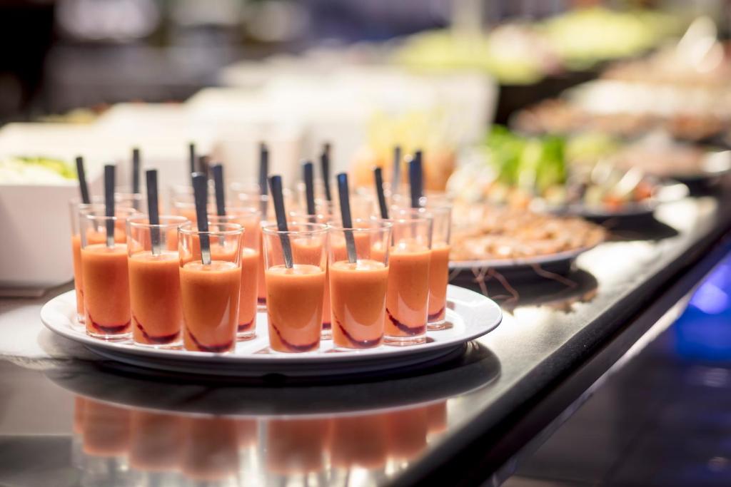 Miniatūras glāzītes ar sarkanu dzērienu uz balta šķīvja, fonā redzami dažādi ēdieni un salāti.