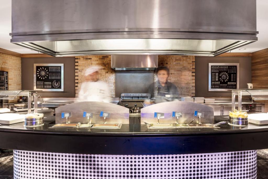 Mūsdienīga virtuves skata ar šefpavāriem, kuri strādā pie atvērtā plīts, un elegantiem virtuves piederumiem uz sienas.