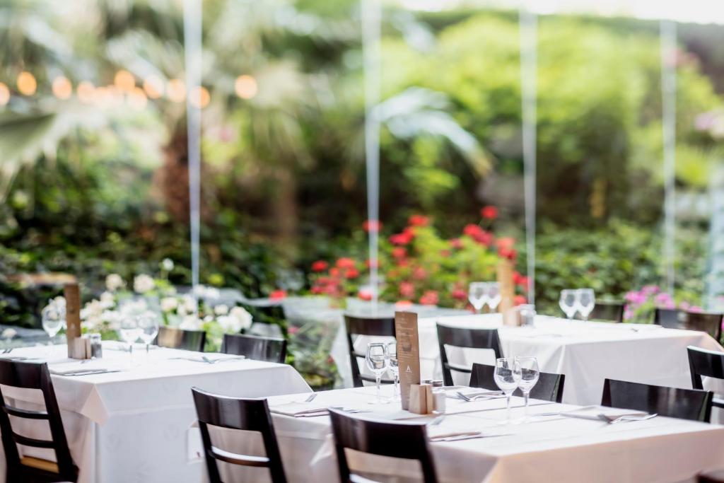 Elegants restorāna interjers ar baltām galdautām un skatu uz zaļo dārzu. Galdi ir sakārtoti ar galda piederumiem un glāzēm, radot patīkamu atmosfēru.