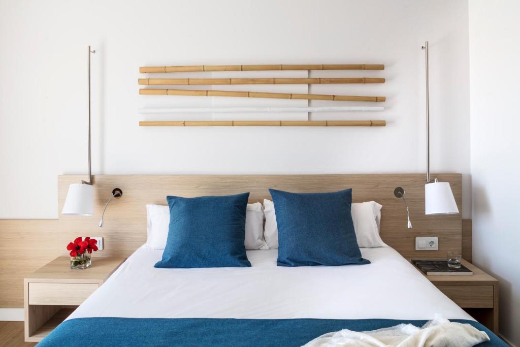 Mūsdienīga guļamistaba ar koka gultas rāmi, zilām spilveniem un minimālisma dizaina apgaismojumu. Uz sienas ir stilizēts bambusa dekors, radot relaksējošu atmosfēru.