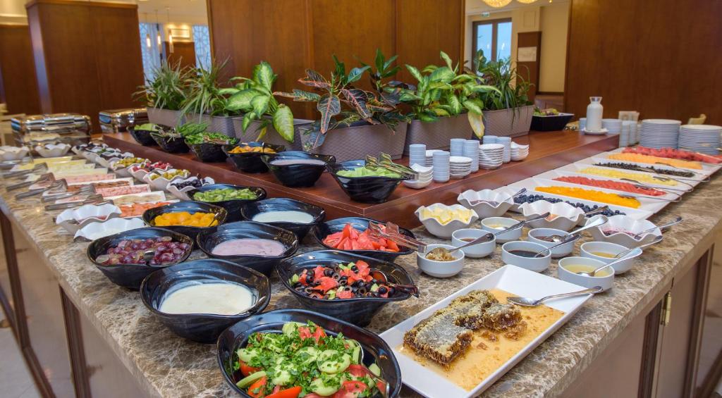 Bagātīgs bufetes galds ar dažādām salātiem, augļiem un desertiem, kas izkārtoti elegantā vidē. Galds rotāts ar augiem un daudziem traukiem, kas pilni ar krāsainiem ēdieniem.