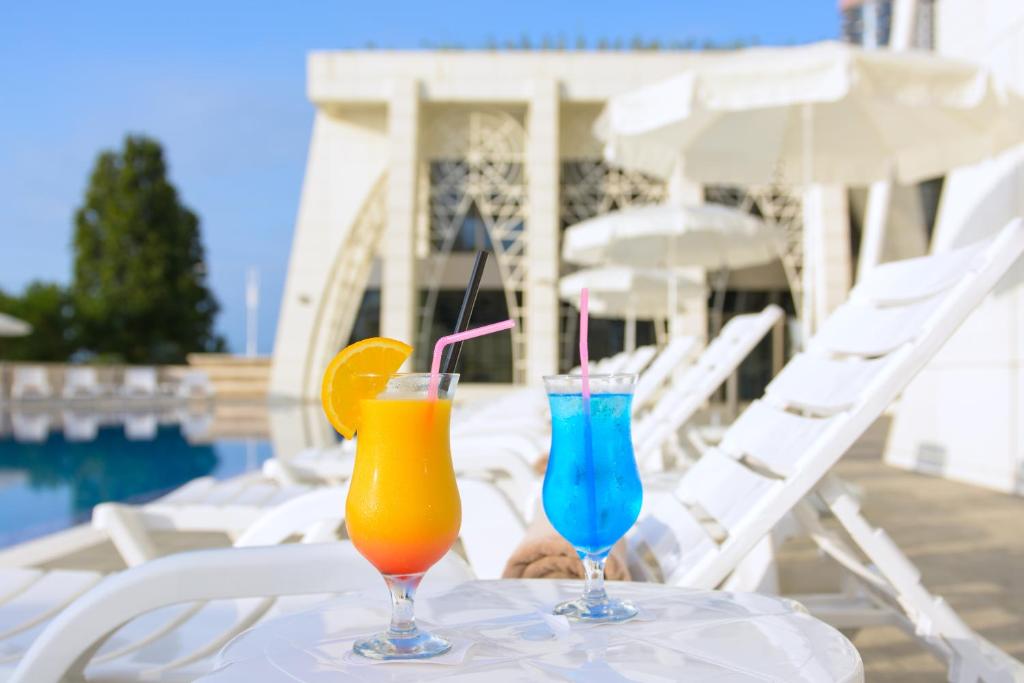 Divi dzērieni ar spilgtām krāsām, oranžs un zils, novietoti uz galdiņa pie baseina, fona redzami sauļošanās krēsli un moderns arhitektūras elements.