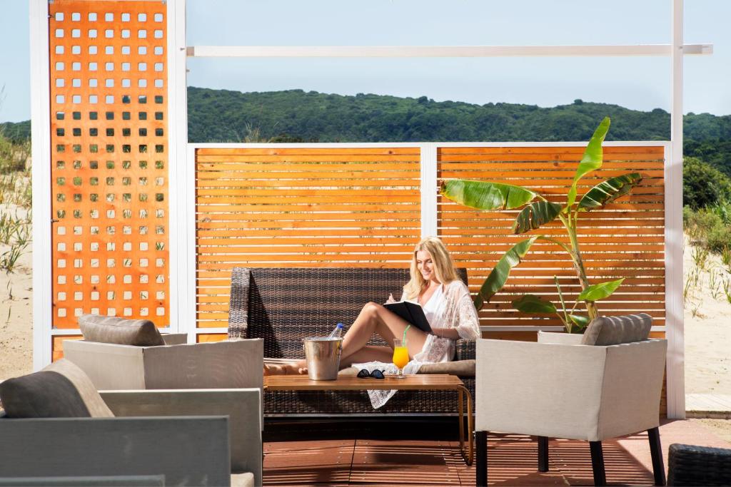 Atpūtas zona pludmalē ar modernām mēbelēm, kur sieviete relaksējas, lasot grāmatu un baudot dzērienu. Fons ar zaļām dabas ainavām un saulainu debesu skatu.