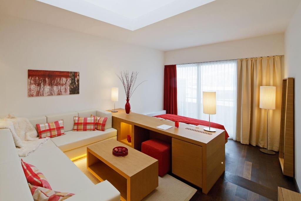 Mūsdienīgs viesistabas interjers ar sarkano un baltu krāsu akcentiem, ietverot ērtu dīvānu, koka galdu un mīkstus spilvenus. Lielas logu aizkari un dabīga apgaismojuma elementi rada patīkamu atmosfēru.