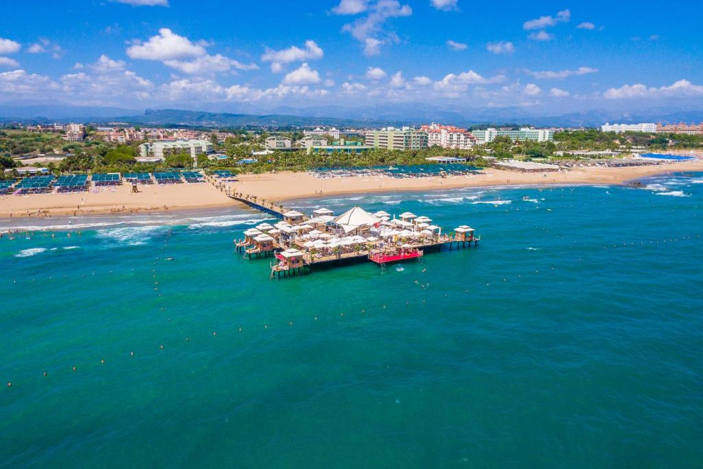 Pludmale ar saulainu debesīm un skaidru ūdeni, uz tās redzams koka piestātne ar atpūtas vietām un cilvēkiem, kas izbauda sauli. Apkārt ir viesnīcas un pludmales krēsli.