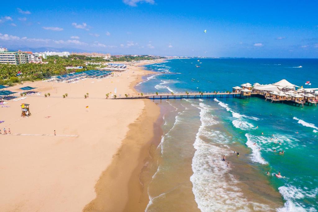 Kāpas piekrastē ar zilu jūru un saulainu debesu fonu, kur pludmalē redzami cilvēki, sauļojoties un peldoties, kā arī koka piestātne ar pludmales restorānu.