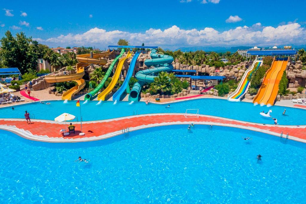 Ūdens atrakciju parks ar krāsainām slidkalniņām un peldbaseinu, kurā redzami apmeklētāji, baudot sauli un ūdens priekus. Pilsētas ainava ar palmām un skaidru debesu fonu.