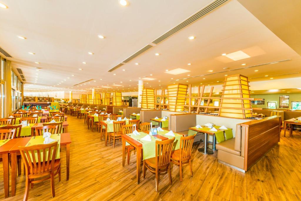 Mūsdienīgs restorāna interjers ar koka galdiem un krēsliem, apgaismots ar siltu gaismu. Uz galdiem ir novietoti zaļi galdauti un salvetes, radot patīkamu atmosfēru.