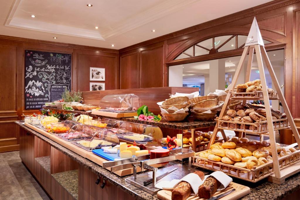 Bagātīgs brokastu bufete ar dažādiem sieriem, maizēm un augļiem, skaisti noformēta koka stendā. Svaigi cepti produkti un krāsaini pasniegšanas trauki rada viesmīlīgu atmosfēru.