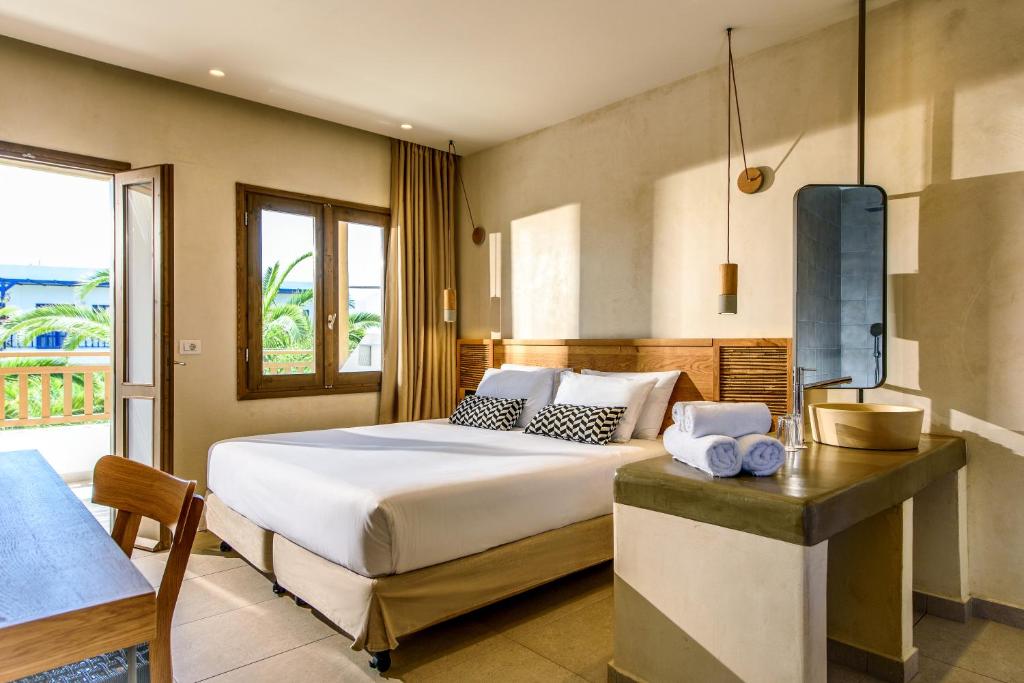 Moderns viesnīcas numurs ar gultu, koka mēbelēm un dabīgu apgaismojumu. Logi paver skatu uz dārzu ar palmām.