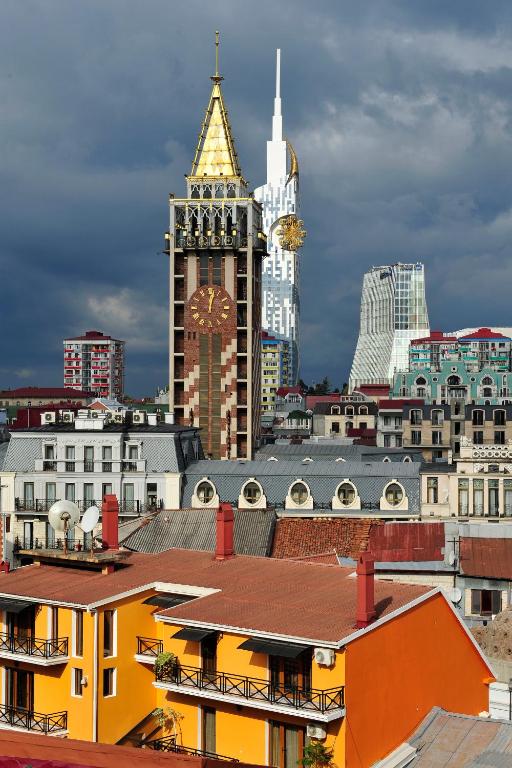 Pilsētas panorāma ar zeltītu pulksteņa torni un mūsdienīgu arhitektūru, kas izceļas uz drūmām debesīm. Tuvumā ir krāsaini māju jumti un dažādas ēku formas.