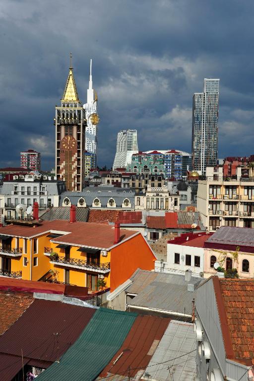 Skats uz pilsētu ar modernām ēkām un vēsturisku torni, kas izceļas ar zeltītu jumtu. Debesis ir apmākušās, piešķirot ainavai dramatisku noskaņu.