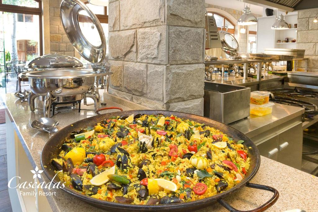 Lielā panna ar krāsainu paelju, kas satur dārzeņus un jūras veltes, ir izvietota pie bufetes galda modernā restorānā. Fonā redzami stainless steel trauki un akmens siena.