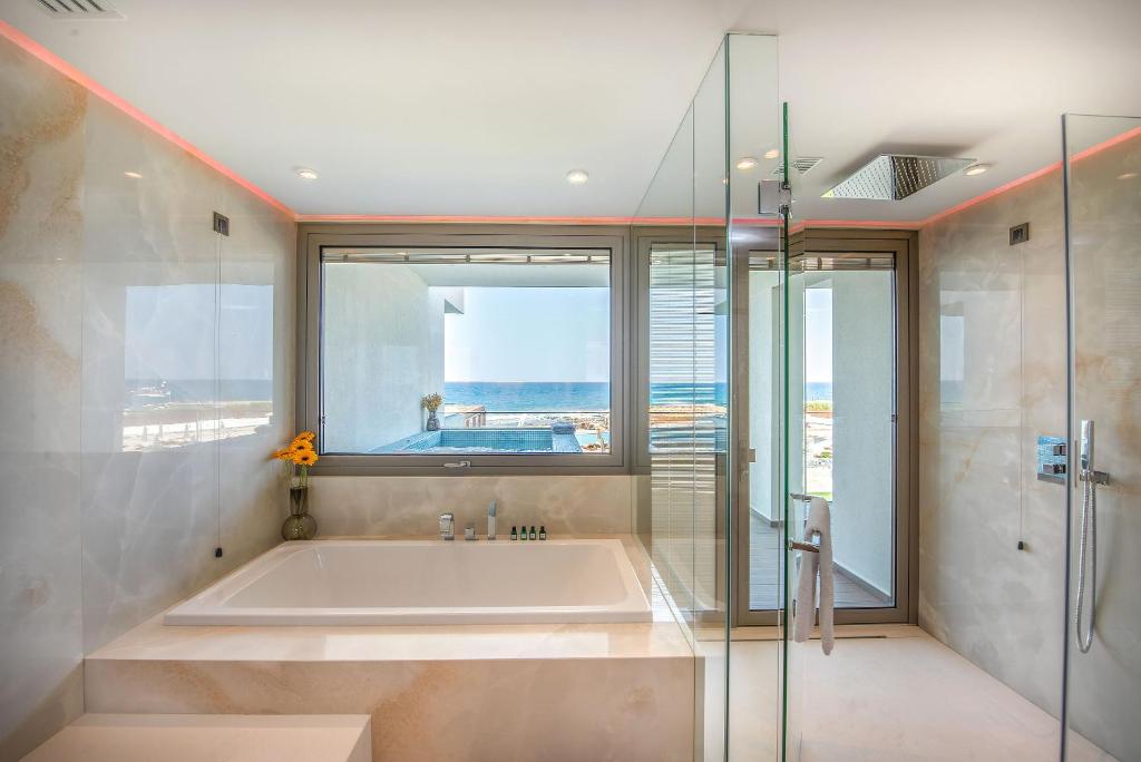 Mūsdienīgs vannas istabas interjers ar skatu uz jūru, kurā ir vannas, duša un lielas logi, kas ļauj ieplūst dabiskajai gaismai. Vannas istabā ir arī dekoratīvas detaļas, piemēram, ziedi un kosmētikas produkti.