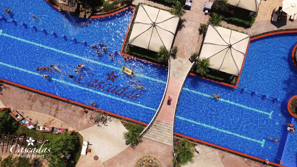 Zilā peldbaseina skats ar cilvēkiem, kas atpūšas un peld, redzami arī sauļošanās krēsli un nojumes. Uz baseina virsmas redzams "Cascadas Family Resort" uzraksts.