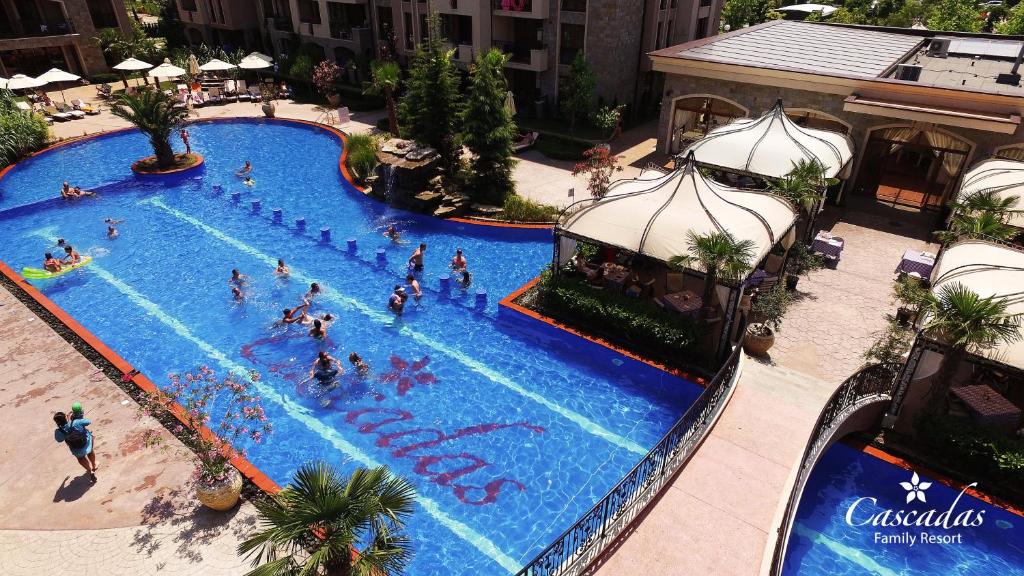 Lielisks baseins ar saulainu atmosfēru, kurā cilvēki atpūšas un izbauda peldēšanu, apņemts ar tropisku zaļumu un komfortablām atpūtas vietām. Cascadas Family Resort logo redzams labajā apakšējā stūrī.