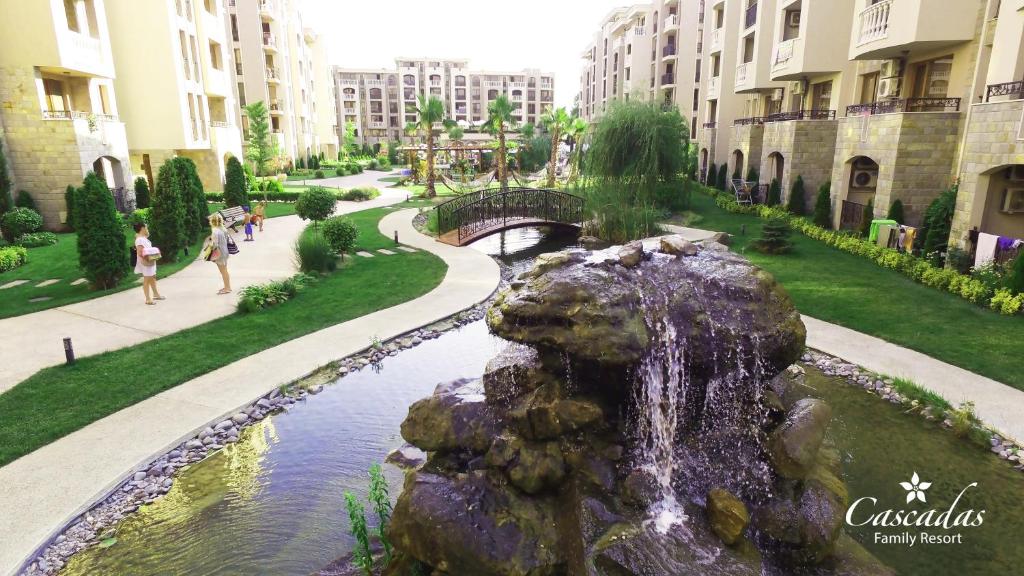 Skaists viesnīcas komplekss ar dārzu un strūklaku, kurā cilvēki pastaigājas pa celiņiem un tiltiem. Āra ainava ar zaļumiem un ūdens elementiem nodrošina mierīgu atmosfēru.